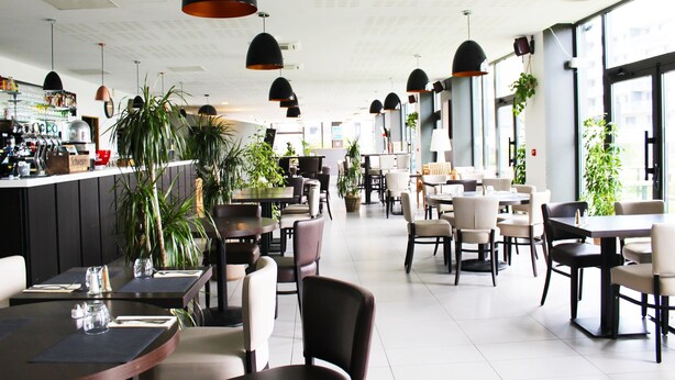 Un interno ristorante moderno con diversi tavoli e sedie, illuminazione a sospensione e ampie finestre che lasciano entrare la luce naturale.