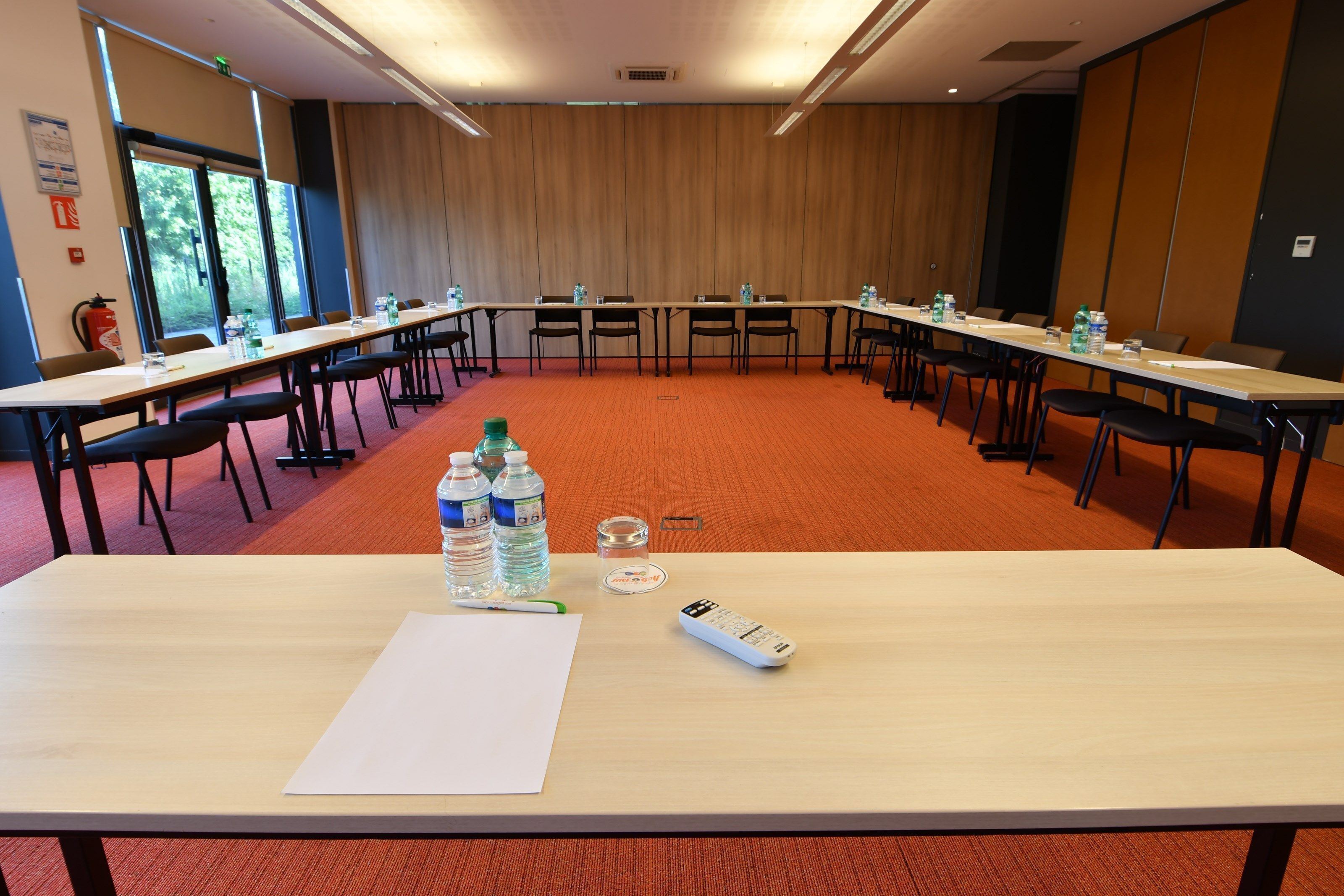 Une salle de conférence équipée de longues tables, de chaises, de bouteilles d’eau, d’une télécommande et d’un tableau blanc.