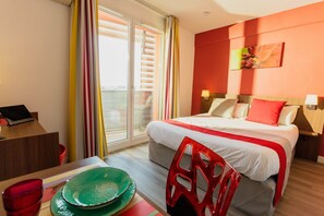 Une chambre d’hôtel avec un lit, un bureau avec un ordinateur portable, une chaise, une table à manger avec un saladier vert et une fenêtre avec des rideaux.