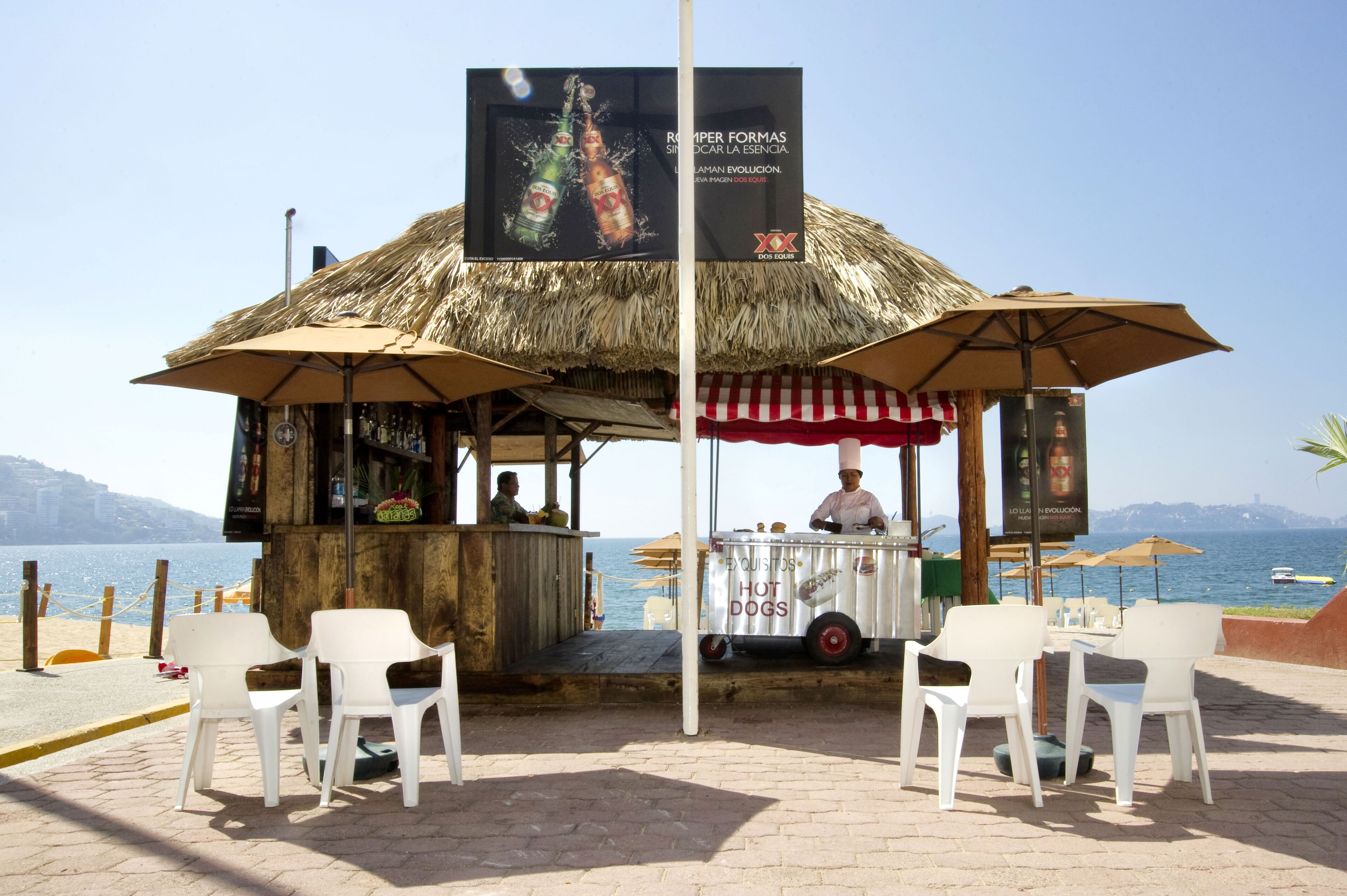 Quiosco frente a la playa con techo de paja, que sirve perritos calientes y ofrece vista al mar.
