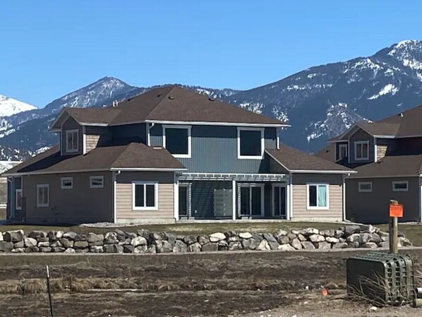 Una casa de dos pisos con base de piedra, un garaje y montañas al fondo.