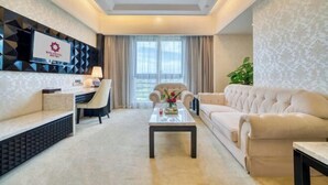 Une chambre d’hôtel moderne, dotée d’un canapé, d’une table basse agrémentée de fleurs, d’un téléviseur fixé au mur et d’une grande fenêtre avec des rideaux.