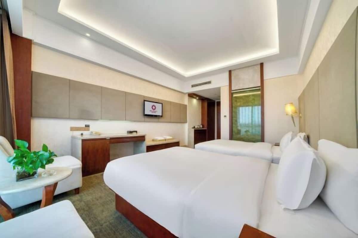 A hotel room with two beds, a desk, a chair, a small table with a plant, and a bathroom visible through a glass door.