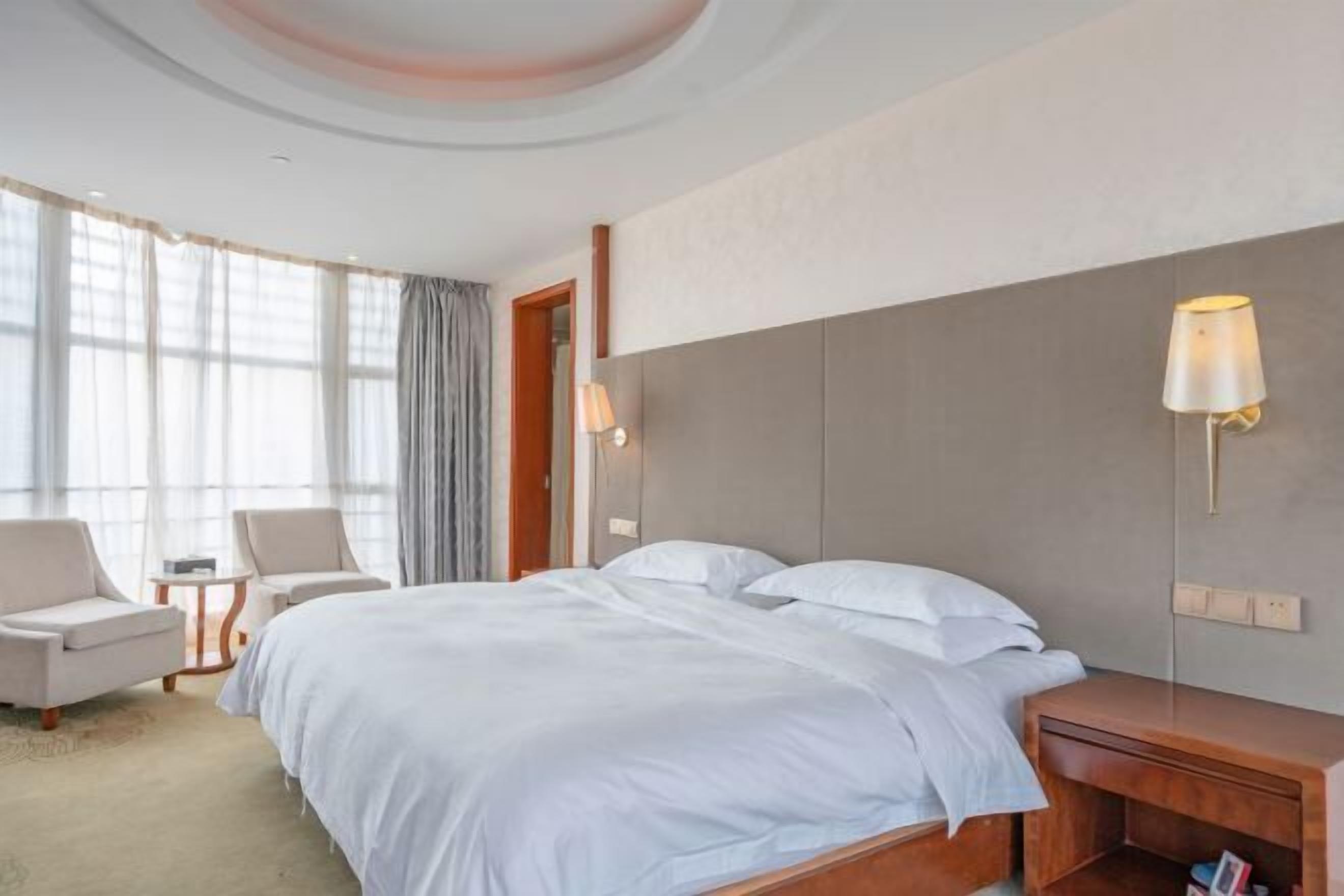 A hotel room with a large bed, two bedside tables, a lamp, and a view of the outdoors through large windows.