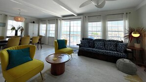 A spacious living room with a large ceiling fan, yellow and blue furniture, and a view of the ocean through large windows. - Waterfront South End Serenity! Bring your boat and your pup, leave the linens!
 (Holly Ridge)