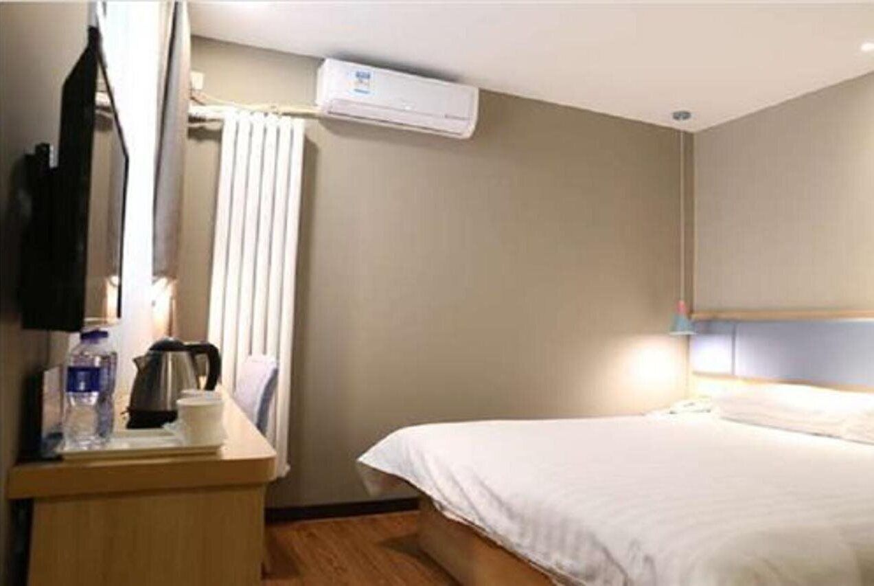 Ein Hotelzimmer mit einem Bett, einem Nachttisch mit Wasserkocher und Flasche, einer Wand-Klimaanlage und einem Fenster mit Vorhang.