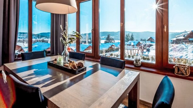 A dining area with a large table, chairs, a hanging lamp, and a view of a snowy landscape through large windows.