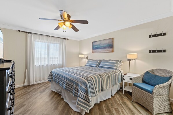 A bedroom with a bed, a chair, a nightstand, a lamp, a ceiling fan, and a painting on the wall.