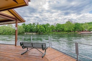A wooden deck with a metal bench overlooking a river, surrounded by lush greenery and a cloudy sky.