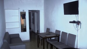 A room with a dining table, chairs, a sofa, a TV mounted on the wall, and a door leading to another room.