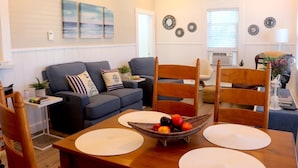 Uma sala de estar com sofá, cadeiras, uma mesa de jantar com jogos americanos, uma fruteira e decorações nas paredes.