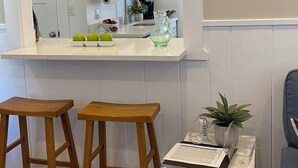 Uma cozinha moderna com armários brancos, bancada e pia. Há dois bancos de madeira, uma mesa pequena com uma planta e um jarro de vidro.