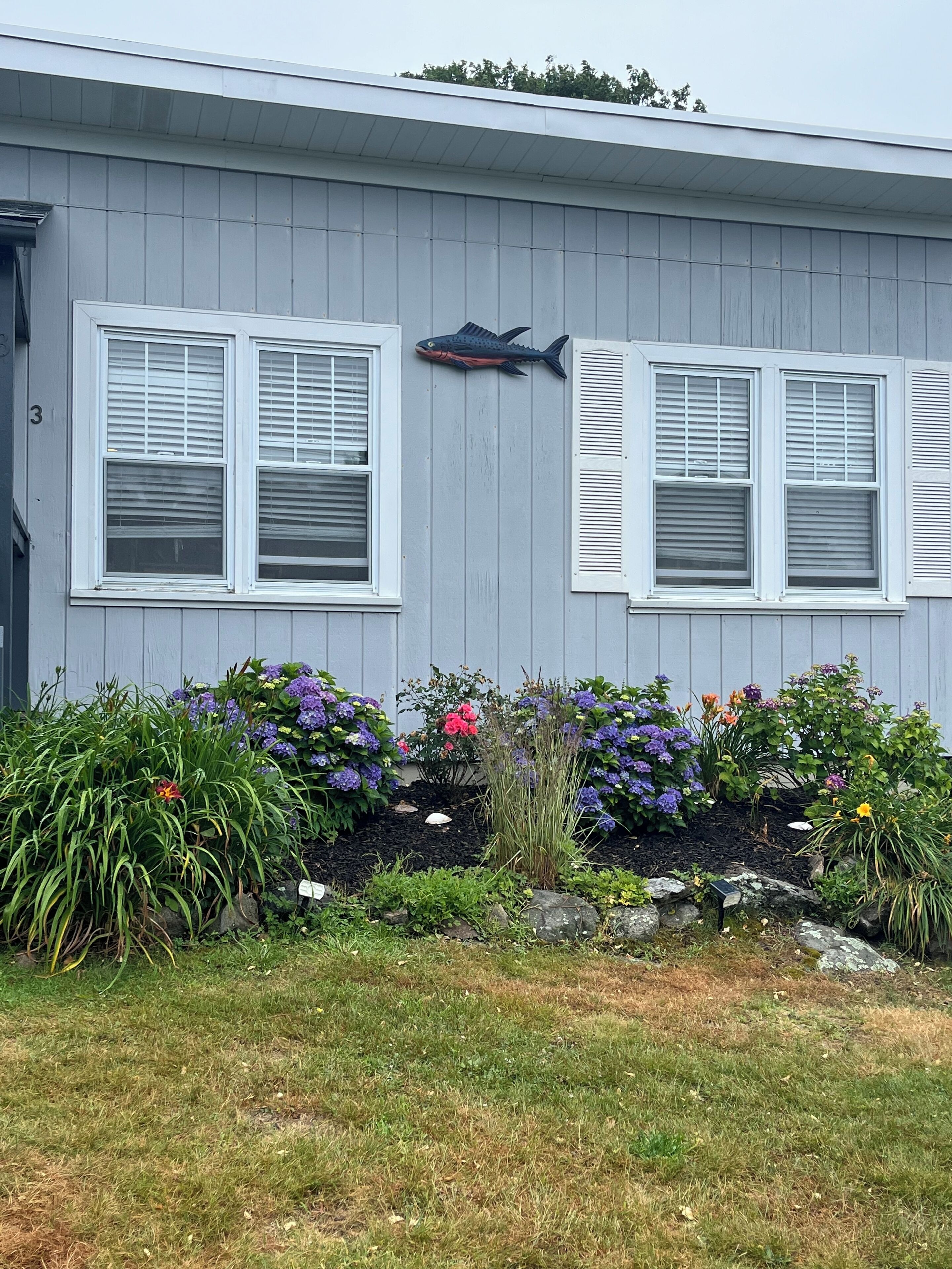 Una casa con dos ventanas, un adorno de pez y un jardín con diversas plantas y flores.
