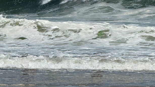 Uma praia com ondas quebrando na costa e dois faróis ao fundo.