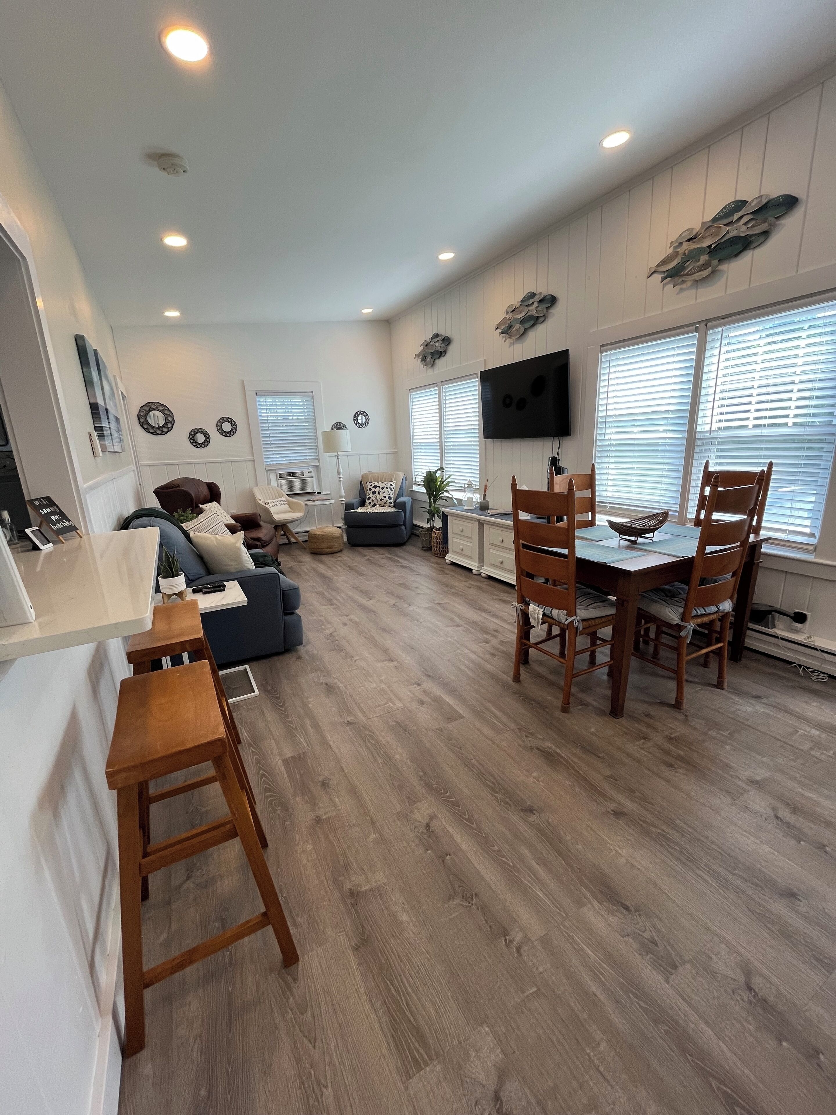 A spacious living area with a wooden floor, a dining table with chairs, a sofa, and a TV mounted on the wall.