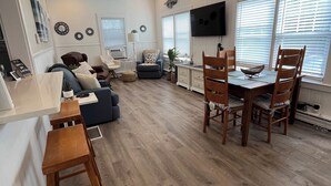 Uma sala de estar ampla, com piso de madeira, uma mesa de jantar com cadeiras, um sofá e uma televisão fixada na parede.