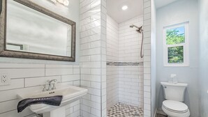 Une salle de bain avec un lavabo blanc, un miroir, une douche avec un sol carrelé et des toilettes.