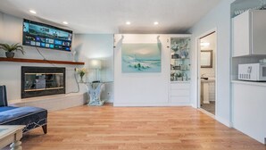 Un vaste espace de vie doté d’un plancher en bois, d’un téléviseur à écran plat fixé au mur, d’un tableau représentant un phare et d’un coin cuisine visible à travers une porte ouverte.