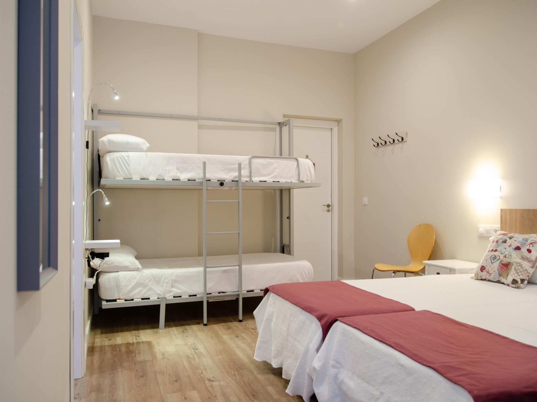 Ein Schlafraum mit Etagenbetten, Schreibtisch und Stuhl, einem Nachttisch und einem Bett mit roter Decke.