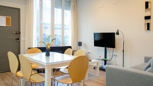 Moderni huone, jossa on lasipöytä, puutuolit, harmaa sohva, litteänäyttöinen televisio ja ikkuna, jossa on sälekaihtimet.