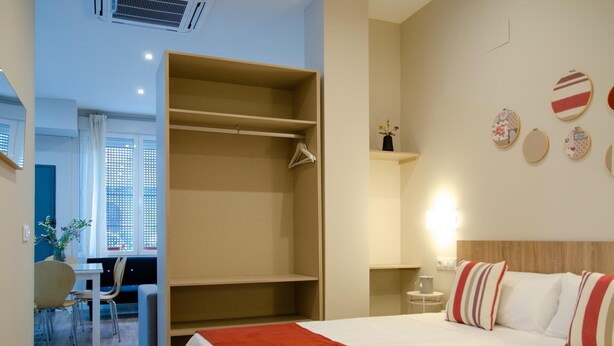 Habitación de hotel con cama, escritorio y silla. Se observa un armario con ropa colgada y un estante con objetos decorativos.