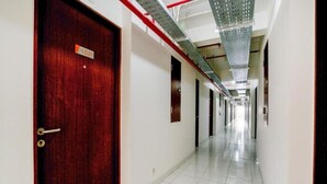 Een lange, smalle hotelgangen met een houten deur aan de linkerkant, witte betegelde vloeren en een metalen plafond met geperforeerde panelen.