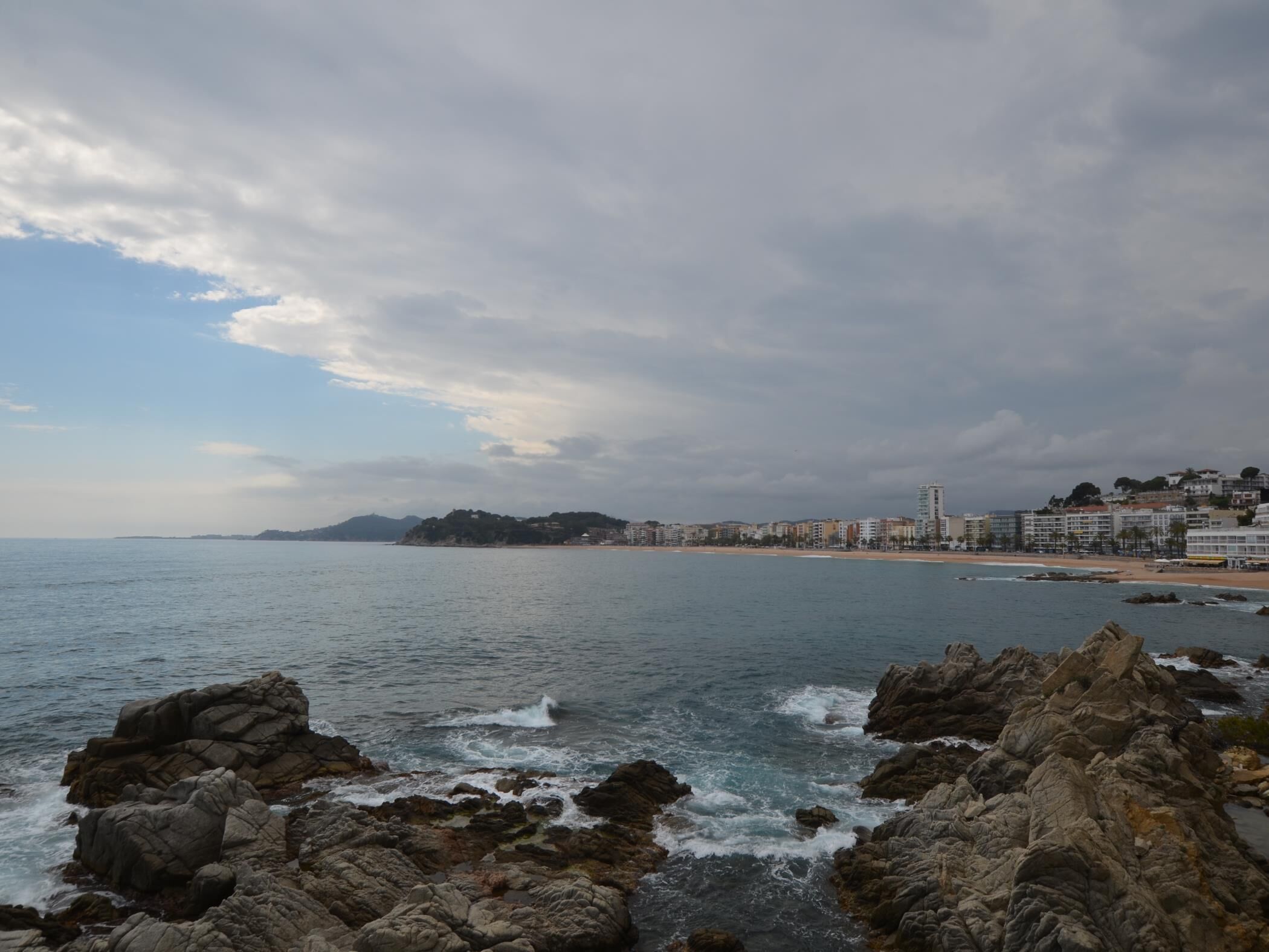 Une vue côtière avec un premier plan rocheux, une mer calme et, au loin, une silhouette urbaine avec des bâtiments.