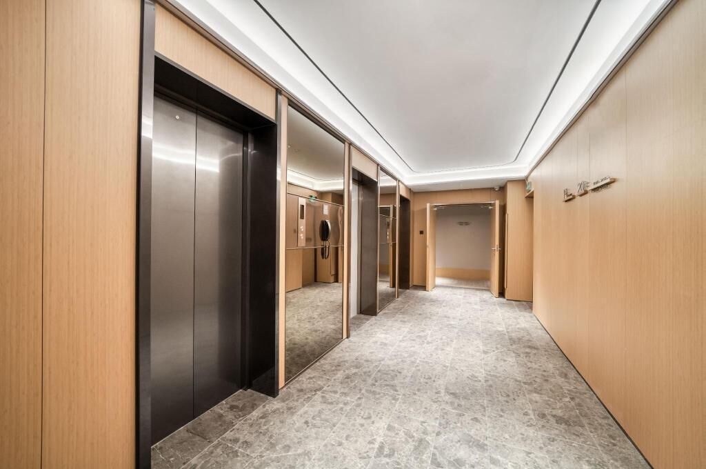A modern hallway with wooden paneling and a marble floor, leading to elevators and rooms.