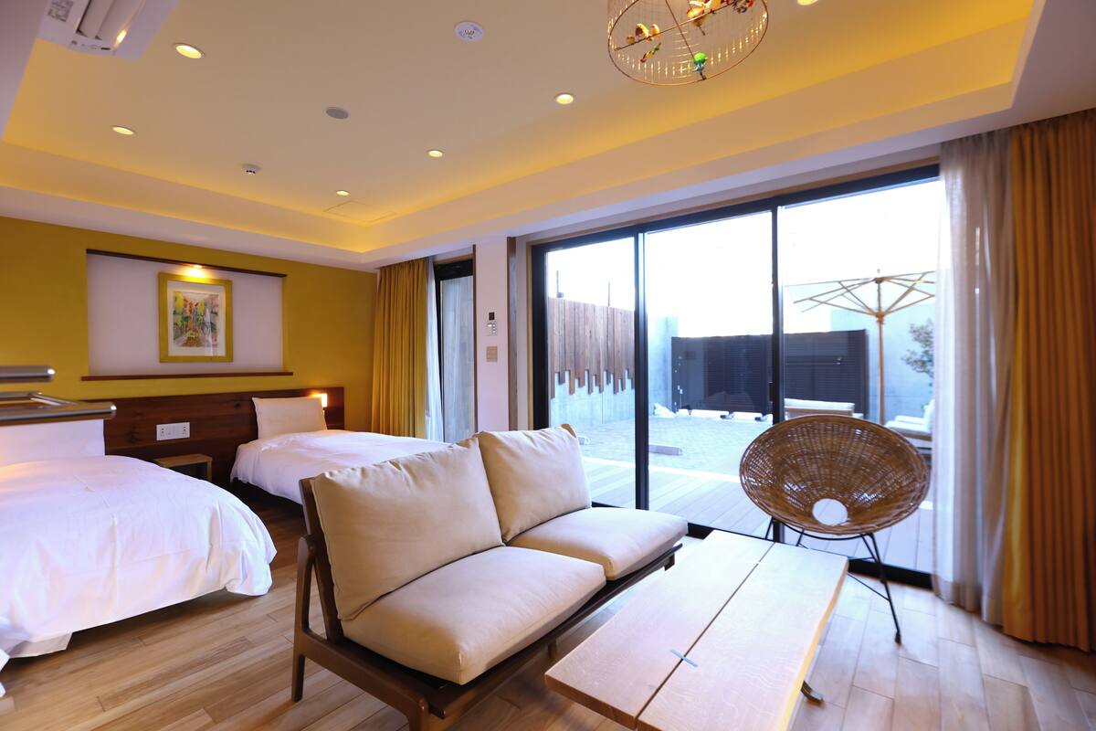 A hotel room with two beds, a sofa, a wicker chair, and a large window with a view.