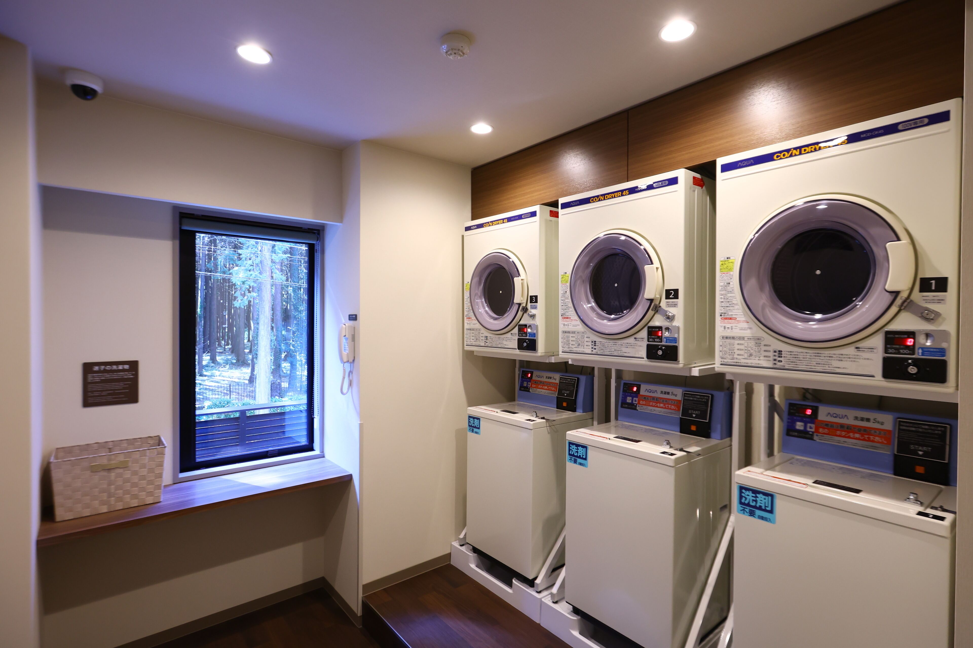 A laundry facility with four washing machines, a window with a view of trees, and a wooden panel on the wall.