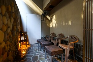Une terrasse dallée de pierres, avec des tables en osier et des chaises en plastique, une porte blanche et un ventilateur fixé au mur.