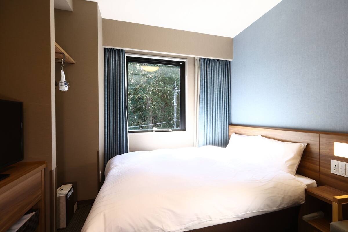 A hotel room with a large bed, a TV on a wooden stand, a window with curtains, and a wall-mounted light.