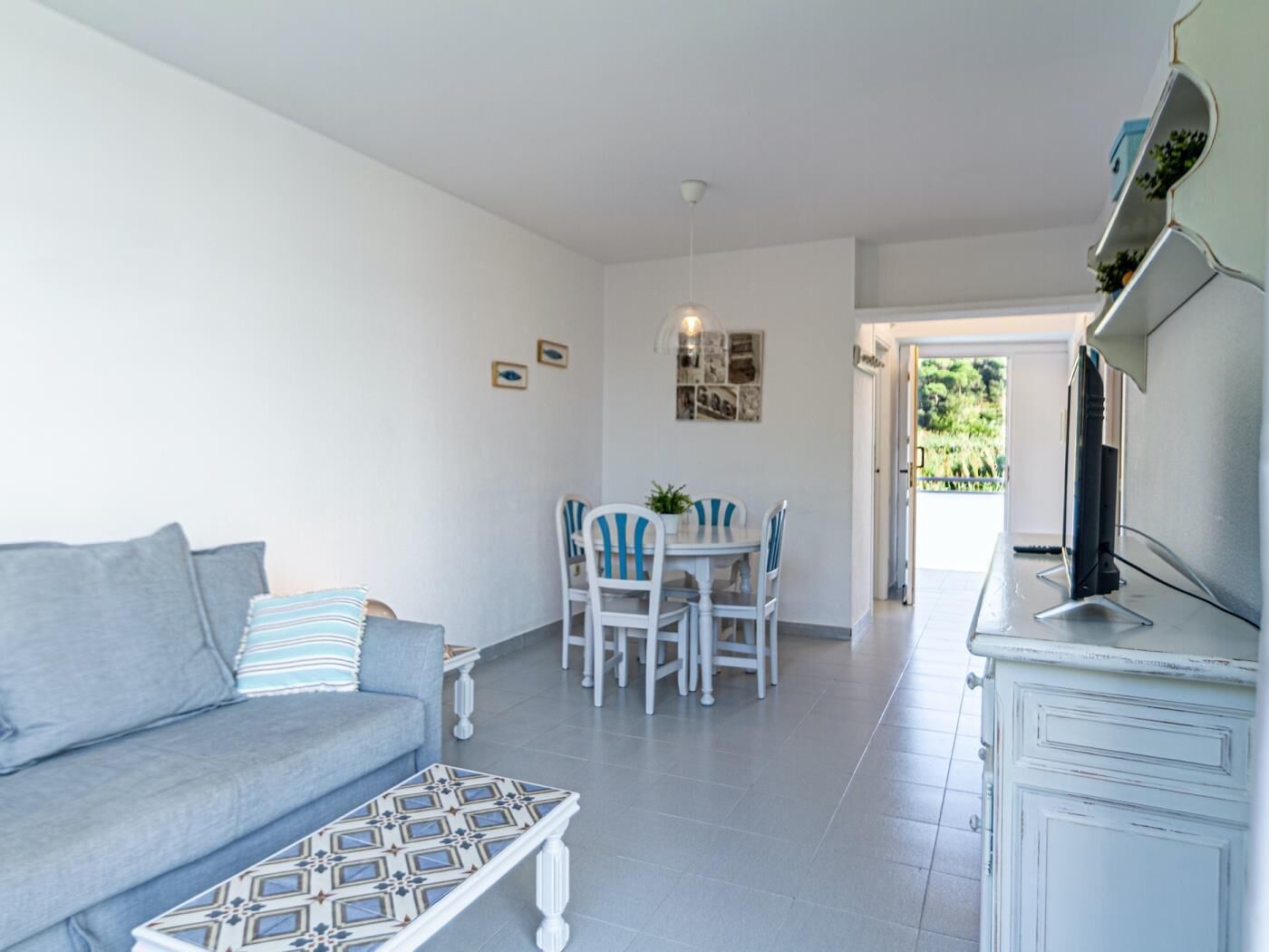 Uma sala de estar com sofá, mesa de centro e área de jantar com mesa e cadeiras. Há uma televisão em um móvel e uma porta que dá para o exterior.