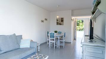 Uma sala de estar com sofá, mesa de centro e área de jantar com mesa e cadeiras. Há uma televisão em um móvel e uma porta que dá para o exterior.