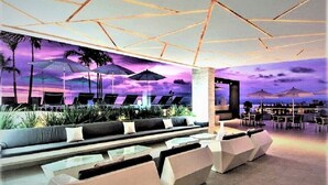 Un área de descanso moderna con un diseño geométrico en el techo, disposición de asientos y un mural que representa un atardecer.