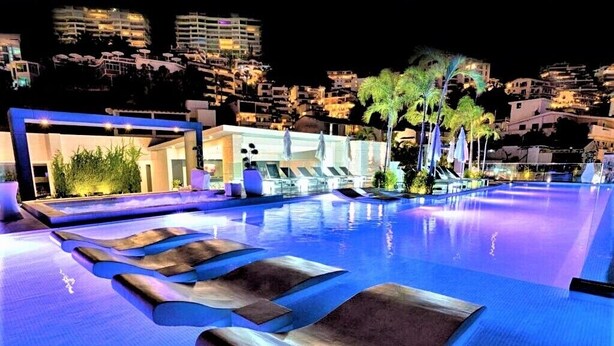 Zona de piscina nocturna con sillones de descanso y vistas a un paisaje urbano al fondo.