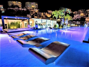 Zona de piscina nocturna amb gandules i vista a un paisatge urbà de fons.
