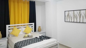 A bedroom with a bed, white nightstands, a bedside table, a wall-mounted artwork, and a ceiling light. - VAMAR LUXURY CONDO- MY PEACE APT (PUERTO PLATA)
