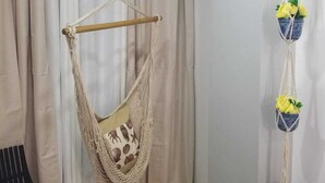 A room with a macramé hanging chair, a small potted plant, and a chair with a floral cushion. - VAMAR LUXURY CONDO- MY PEACE APT (PUERTO PLATA)