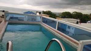 A swimming pool with a glass barrier, a metal handrail, and a view of the sea and buildings in the background. - VAMAR LUXURY CONDO- MY PEACE APT (PUERTO PLATA)