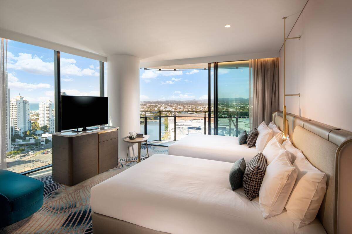 A modern hotel room with a large bed, a TV, a small desk, and a balcony with a city view.