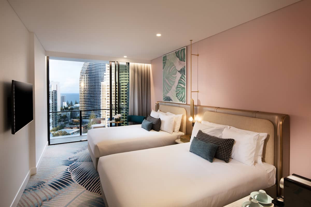 A modern hotel room with two beds, a large window with a city view, a flat-screen TV, and a balcony with a view of high-rise buildings.