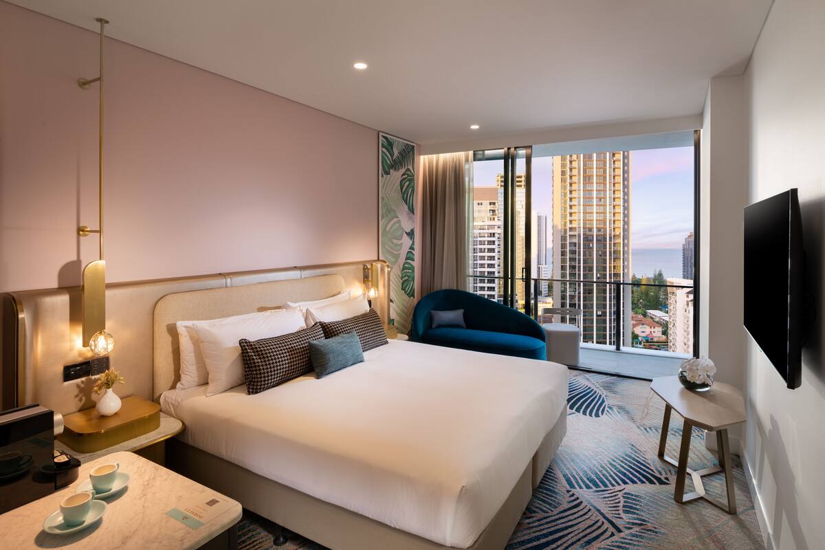 A modern hotel room with a large bed, a sitting area with a blue chair, a flat-screen TV, and a city view through large windows.