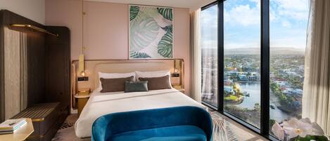 A modern hotel room with a large bed, a blue sofa, a view of a cityscape through large windows, and a tropical-themed artwork on the wall.