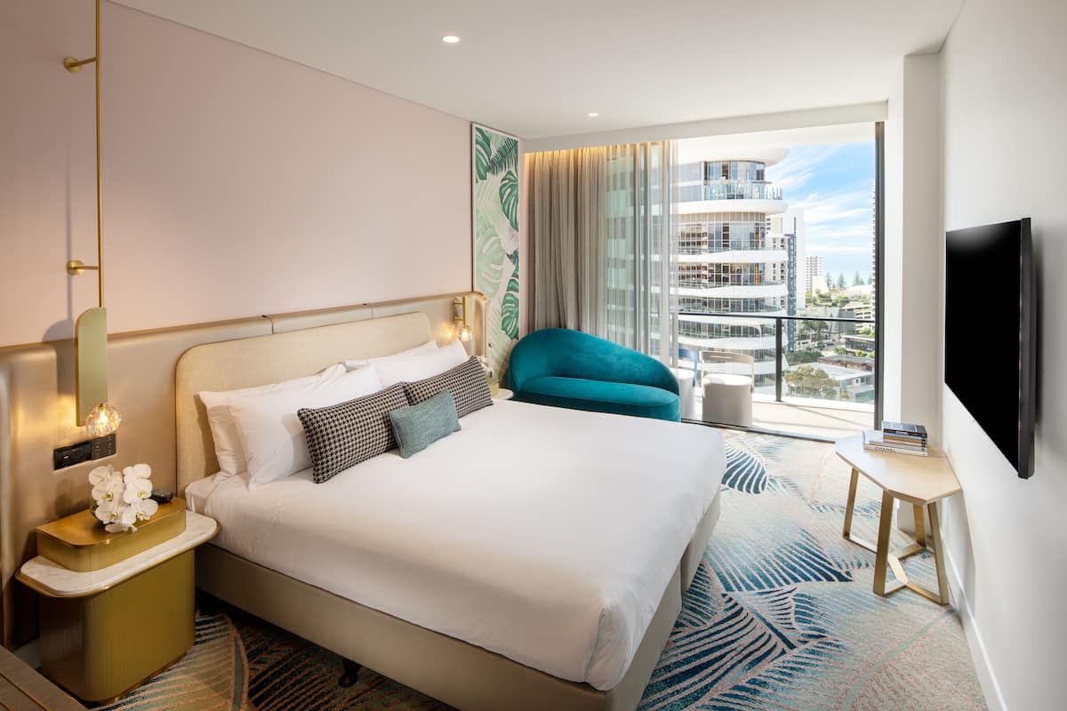 A modern hotel room with a large bed, a teal armchair, a small table, and a TV.