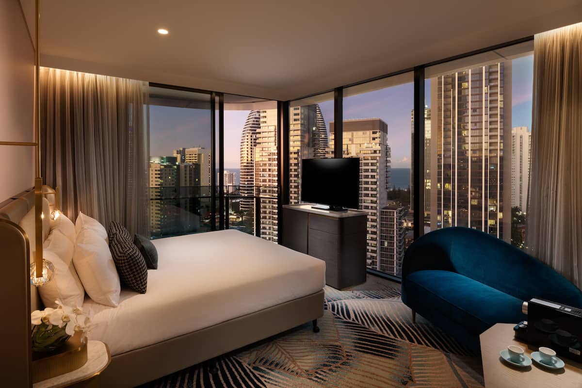 A modern hotel room with a large bed, a sofa, a TV, and a city view.
