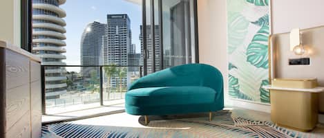 A modern hotel room with a teal armchair, a large window with a city view, a patterned rug, and a minimalist side table.