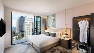 Una habitación de hotel moderna con una cama grande, vistas a la ciudad, un armario con albornoces y una mesita de noche con lámpara.