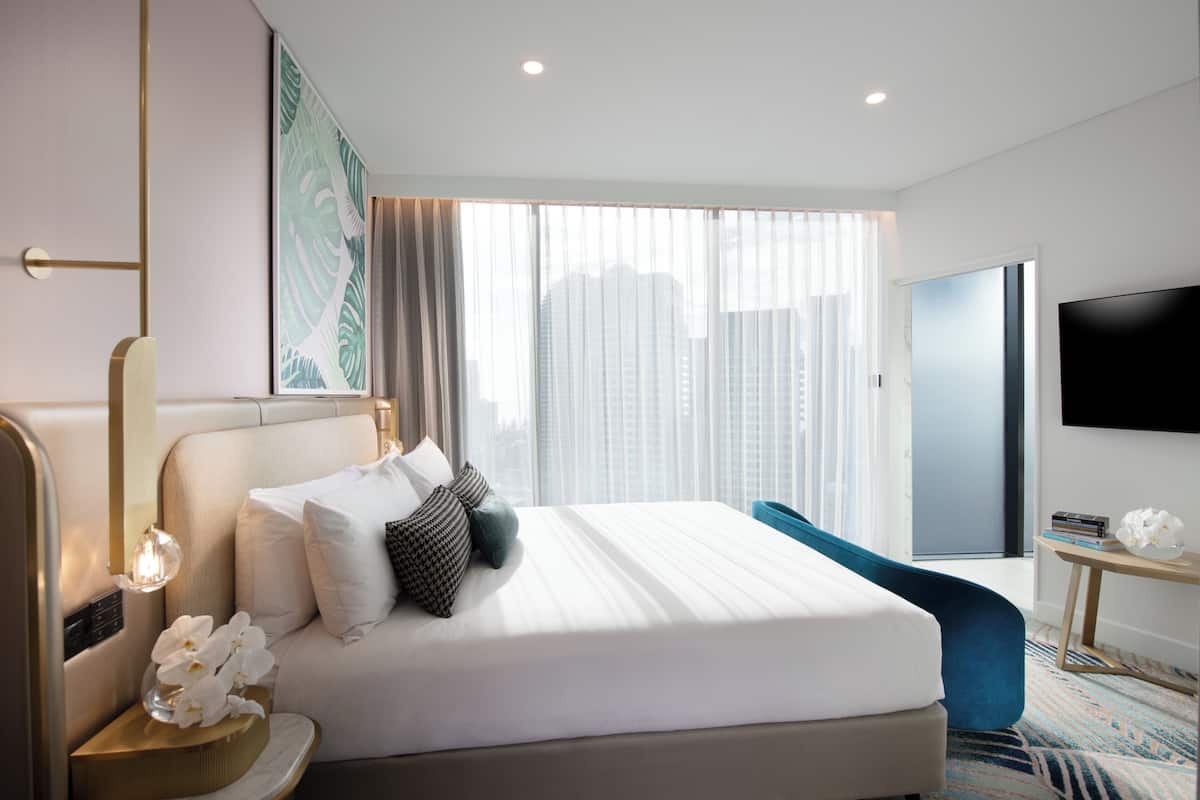 A modern hotel room with a large bed, a TV, a nightstand with a lamp, and a view of the city through the window.