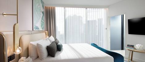 A modern hotel room with a large bed, a TV, a nightstand with a lamp, and a view of the city through the window.